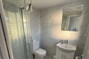 A bathroom with a glass shower enclosure, a toilet, a sink with a mirror above it, and marble-patterned walls. - Near Heathrow - Studio - WiFi - Parking - Sleeps 2 (Uxbridge)