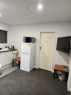 A compact kitchen with a refrigerator, microwave, toaster, and kettle. There is a small wooden table and a TV mounted on the wall.