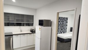 A compact kitchen with a refrigerator, toaster, and dishwasher, leading to a bedroom with a bed and a patterned curtain.