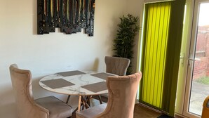 A dining area with a round table, four chairs, a wall-mounted art piece, a potted plant, and a sliding glass door with a green curtain. - House Sleeps 8 with WiFi (Darthford)