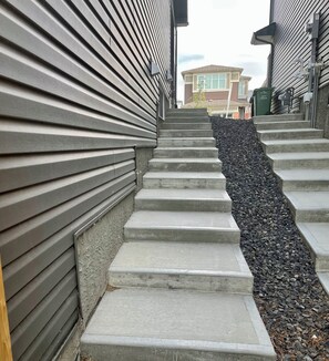 Un escalier en béton, avec un sentier étroit et rocheux à côté, bordé de bâtiments aux bardages horizontaux.