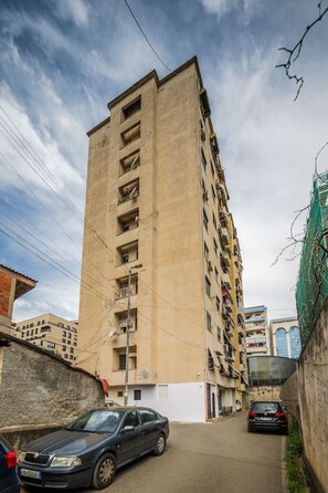 A tall, beige building with multiple balconies, a narrow street, and parked cars. - City Vibes Studio Tirana Center (Tirana)