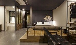Quarto de hotel moderno com cama, um banco de madeira, uma pequena mesa e um banheiro visível por uma porta aberta.