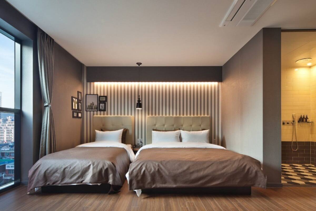 Quarto de hotel com duas camas, vista para a cidade, área de chuveiro moderna e uma janela grande com cortinas.