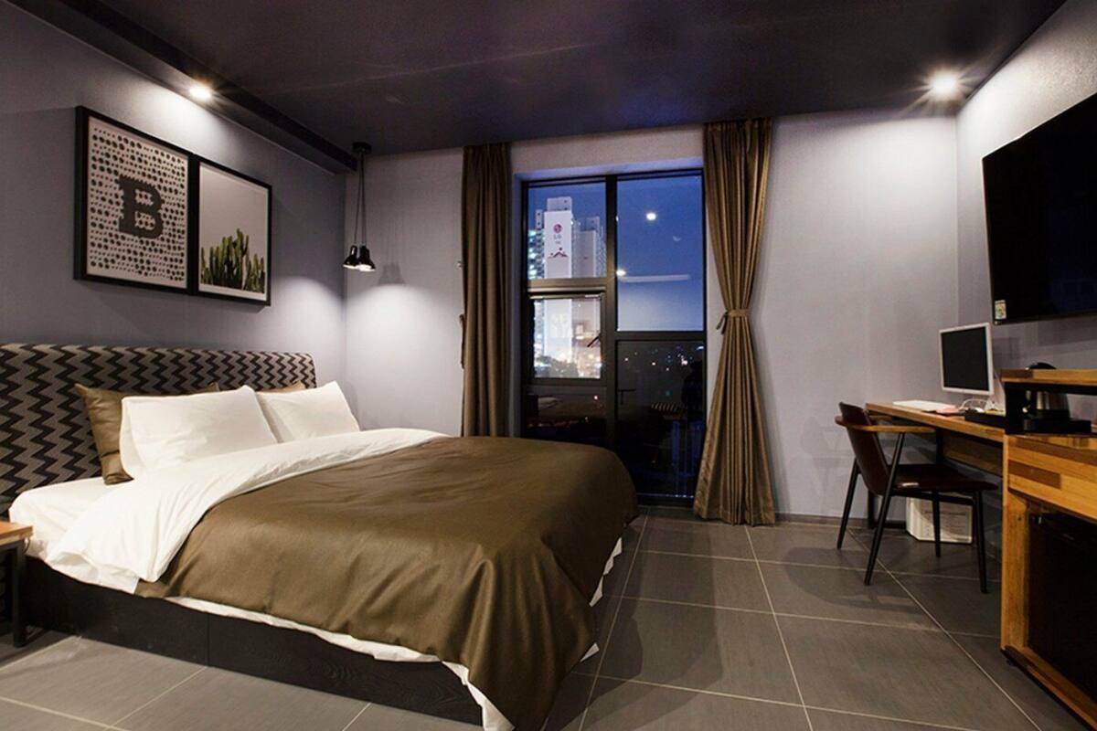 Quarto de hotel moderno com uma cama grande, uma escrivaninha com computador, televisão e varanda com vista para a cidade.