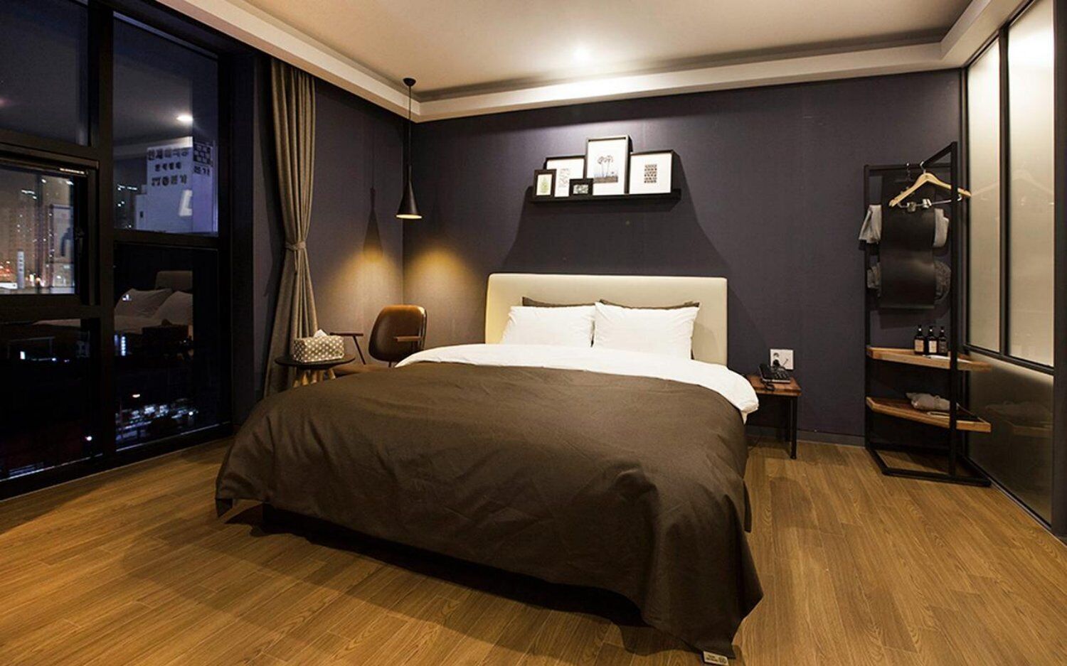A modern hotel room with a large bed, a desk, a chair, a nightstand, and a wardrobe.