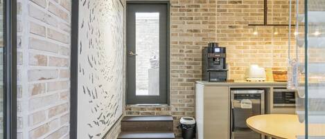 A kitchen with a brick wall, a coffee machine, a refrigerator, and a dining area with a table and bench.