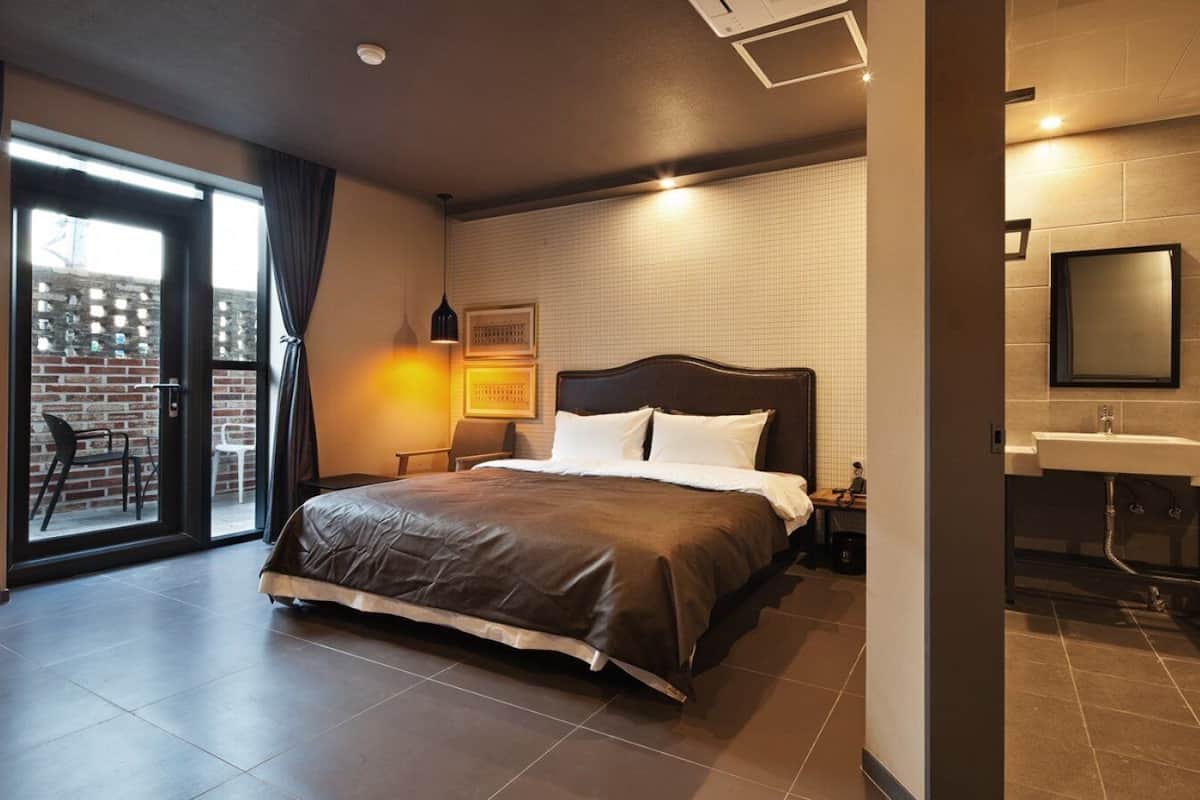 Quarto de hotel moderno com uma cama grande, banheiro com pia e espelho, e vista através de uma porta de vidro para área externa.