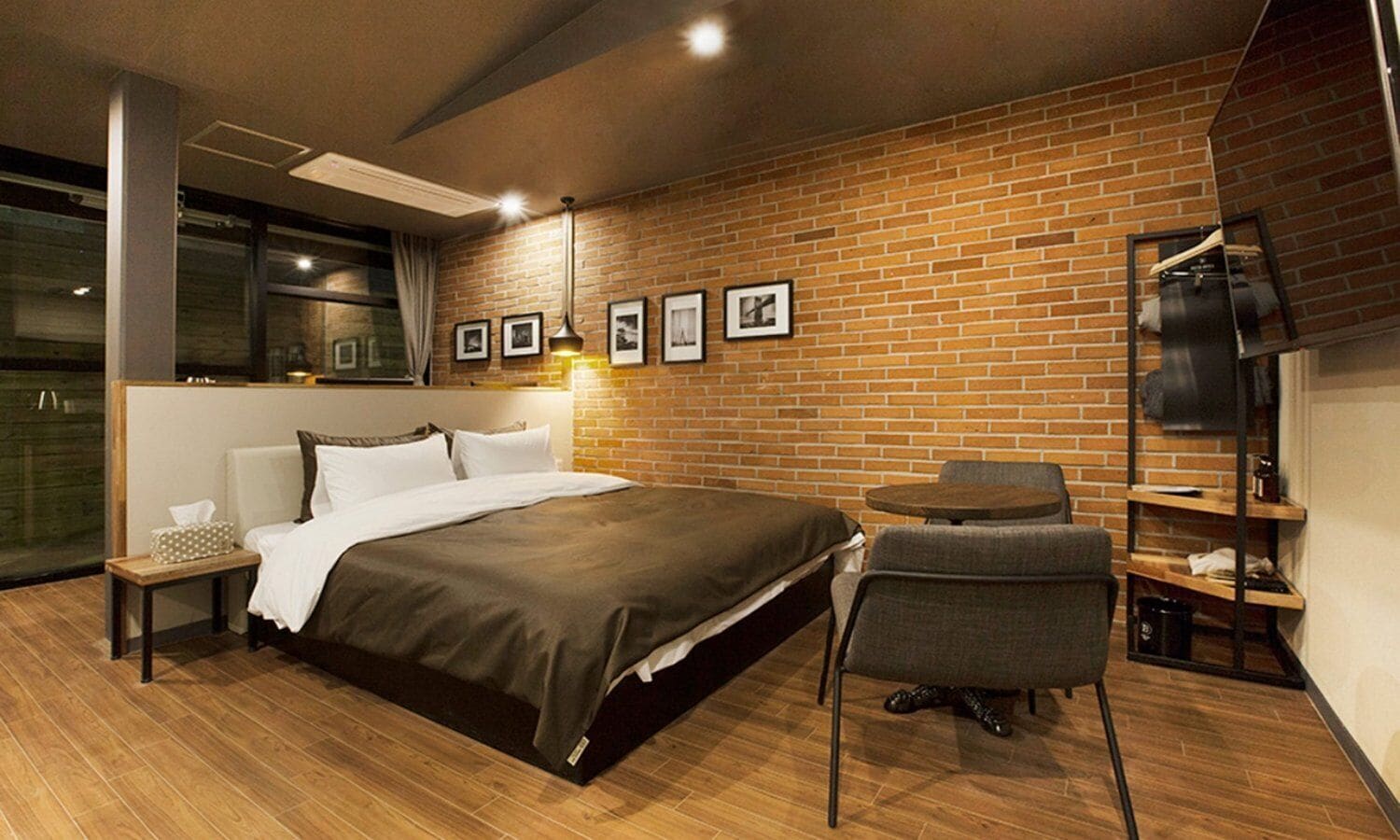 A modern hotel room with a brick wall, a bed, a chair, a small table, and a TV on a shelf.