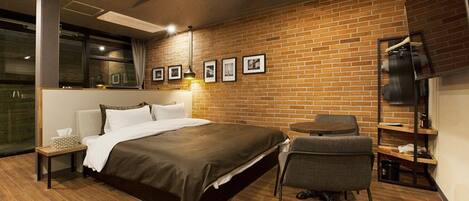 A modern hotel room with a brick wall, a bed, a chair, a small table, and a TV on a shelf.