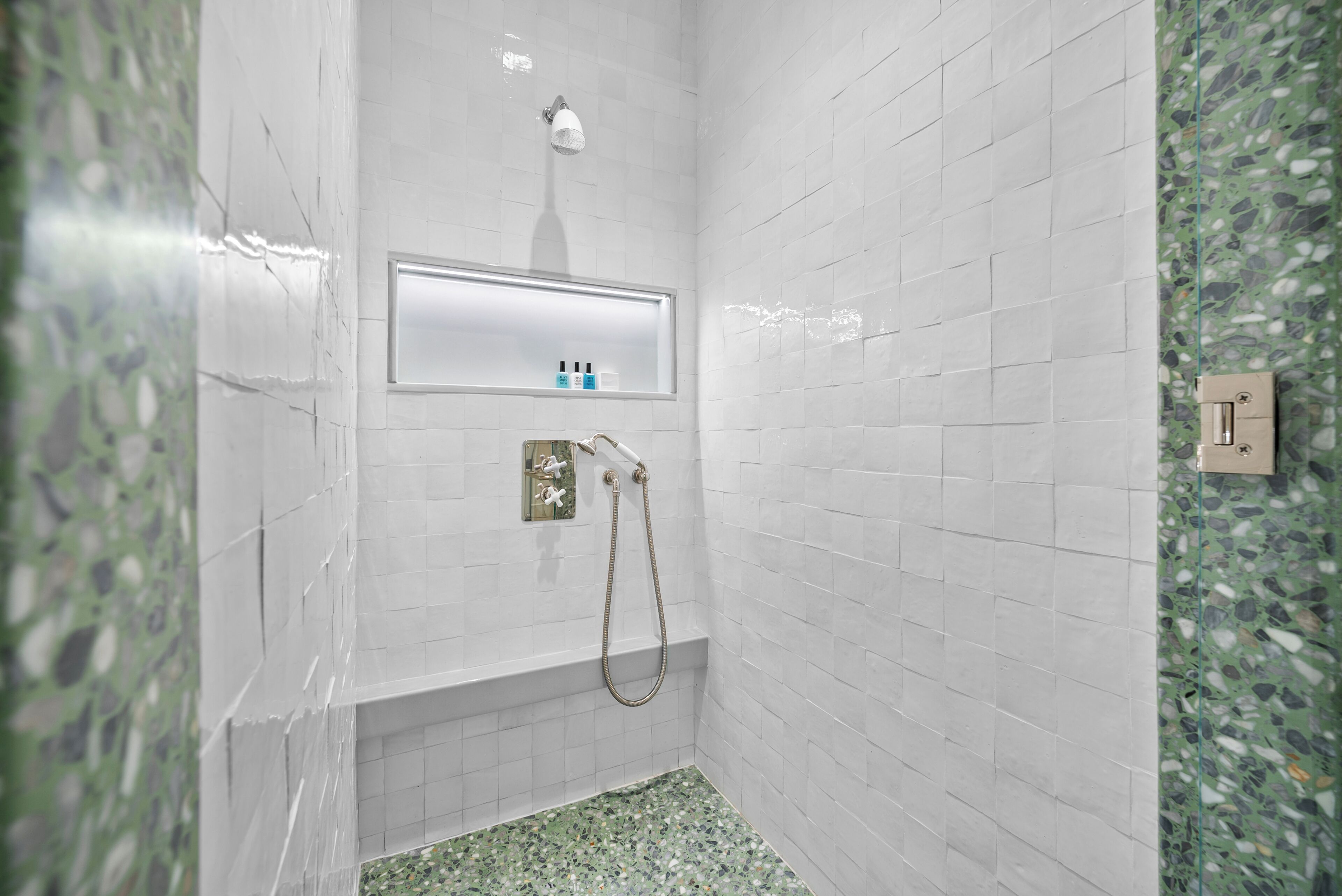 Un baño con paredes de azulejos blancos, piso de mosaico verde, cabezal de ducha de mano e iluminación empotrada.