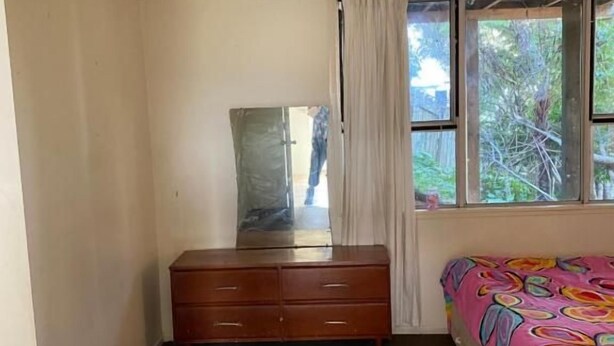 Une chambre avec une commode en bois, un lit recouvert d’une courtepointe colorée, une fenêtre avec des rideaux et un miroir accroché au mur.