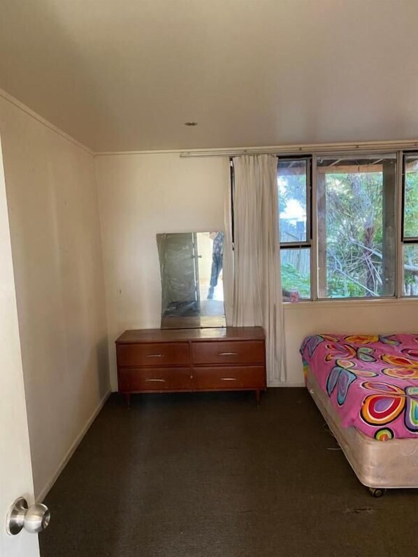 A bedroom with a wooden dresser, a bed with a colorful bedspread, a window with curtains, and a mirror on the wall.