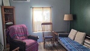 Ein Zimmer mit Deckenventilator, einem fenster mit Vorhängen, einem Holzschaukelstuhl, einem Sessel mit Decke, einem Bett mit gemusterten Betttextilien und einer Lampe.