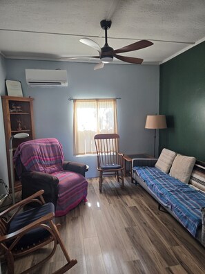 Ein Zimmer mit Deckenventilator, einem fenster mit Vorhängen, einem Holzschaukelstuhl, einem Sessel mit Decke, einem Bett mit gemusterten Betttextilien und einer Lampe.