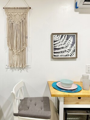 A dining area with a macrame wall hanging, a framed botanical print, a wooden table with a blue placemat, a white chair, and a kitchen area with an oven. - Clean GATED BeachBoho Private Tiny Suite AC/Heat WasherDryer by LAX+Beach+SoFi (Inglewood)