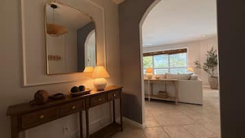 A hallway with a wooden console table, a mirror, and a lamp. There is a doorway leading to a living area with a sofa and a window.