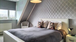 A modern bedroom with a sloped ceiling, a large bed with patterned pillows, a chair, and a bedside lamp.