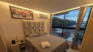 A bedroom with a large bed, a nightstand, a lamp, an air conditioner, a painting, and a view of a cityscape through a window. - Comfortable private apartment near the Tennis Club in Cali salsa city with AC. (Cali)