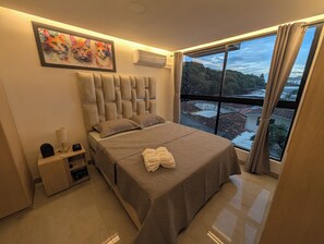 Een slaapkamer met een groot bed, een nachtkastje, een lamp, een airconditioner, een schilderij en uitzicht op een stadsgezicht door een raam.