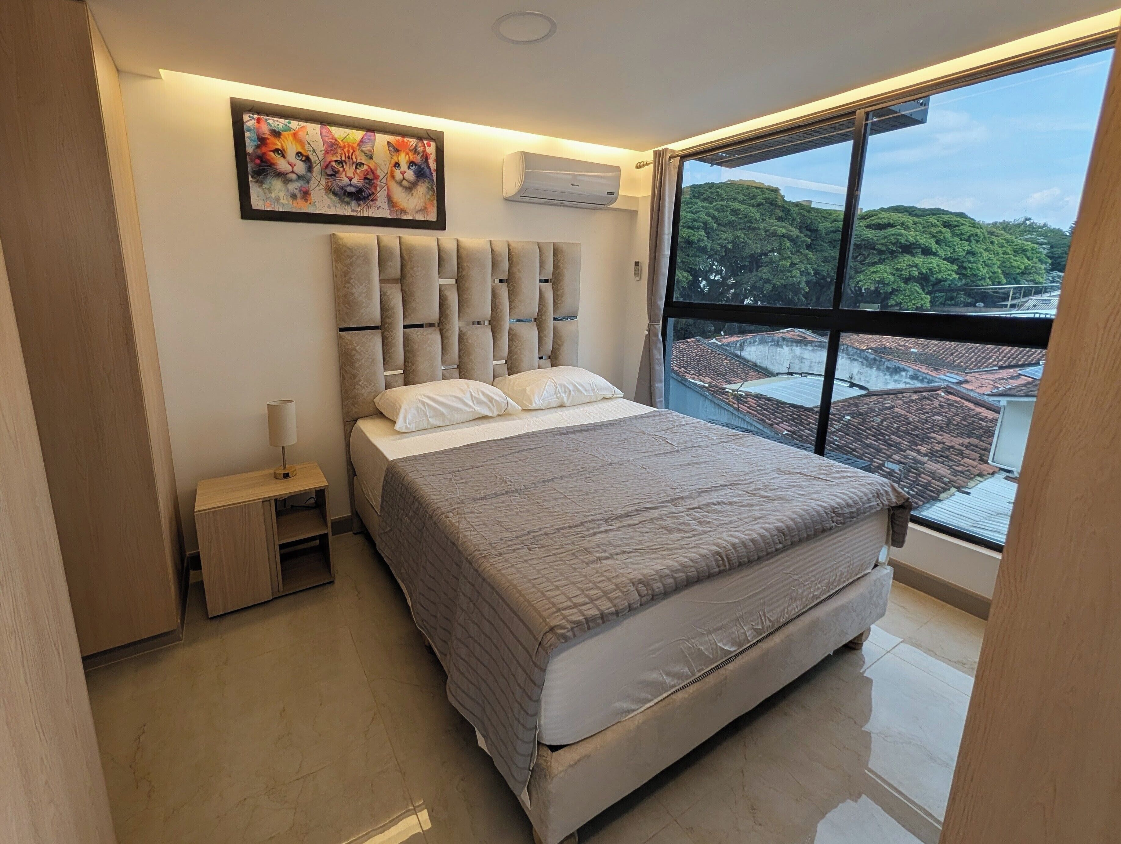 A modern bedroom with a large bed, a nightstand, a wall-mounted air conditioner, and a large window with a city view.