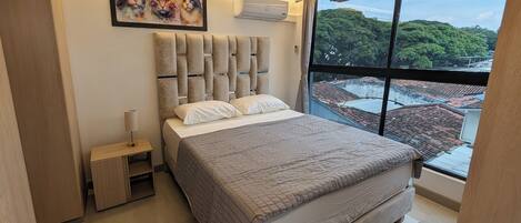 A modern bedroom with a large bed, a nightstand, a wall-mounted air conditioner, and a large window with a city view.