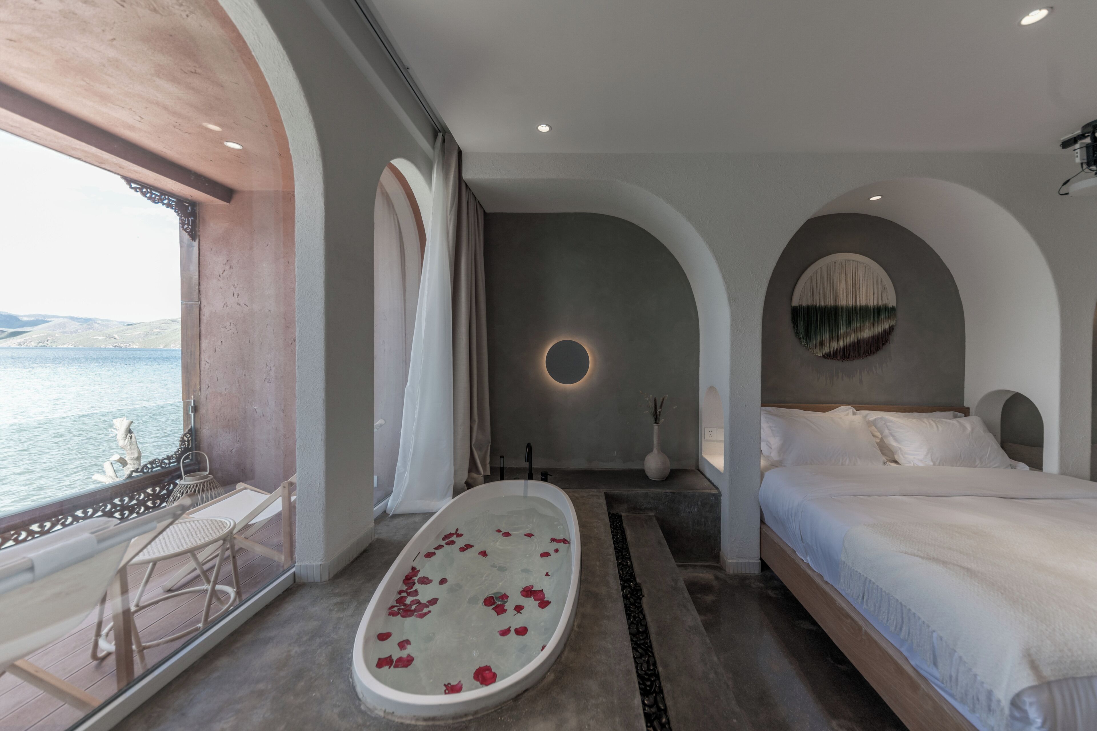 Un dormitorio con una cama grande, una bañera con pétalos de rosa, vistas al mar y un balcón con mesa y sillas.