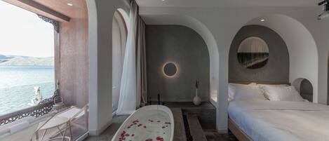 Un dormitorio con una cama grande, una bañera con pétalos de rosa, vistas al mar y un balcón con mesa y sillas.