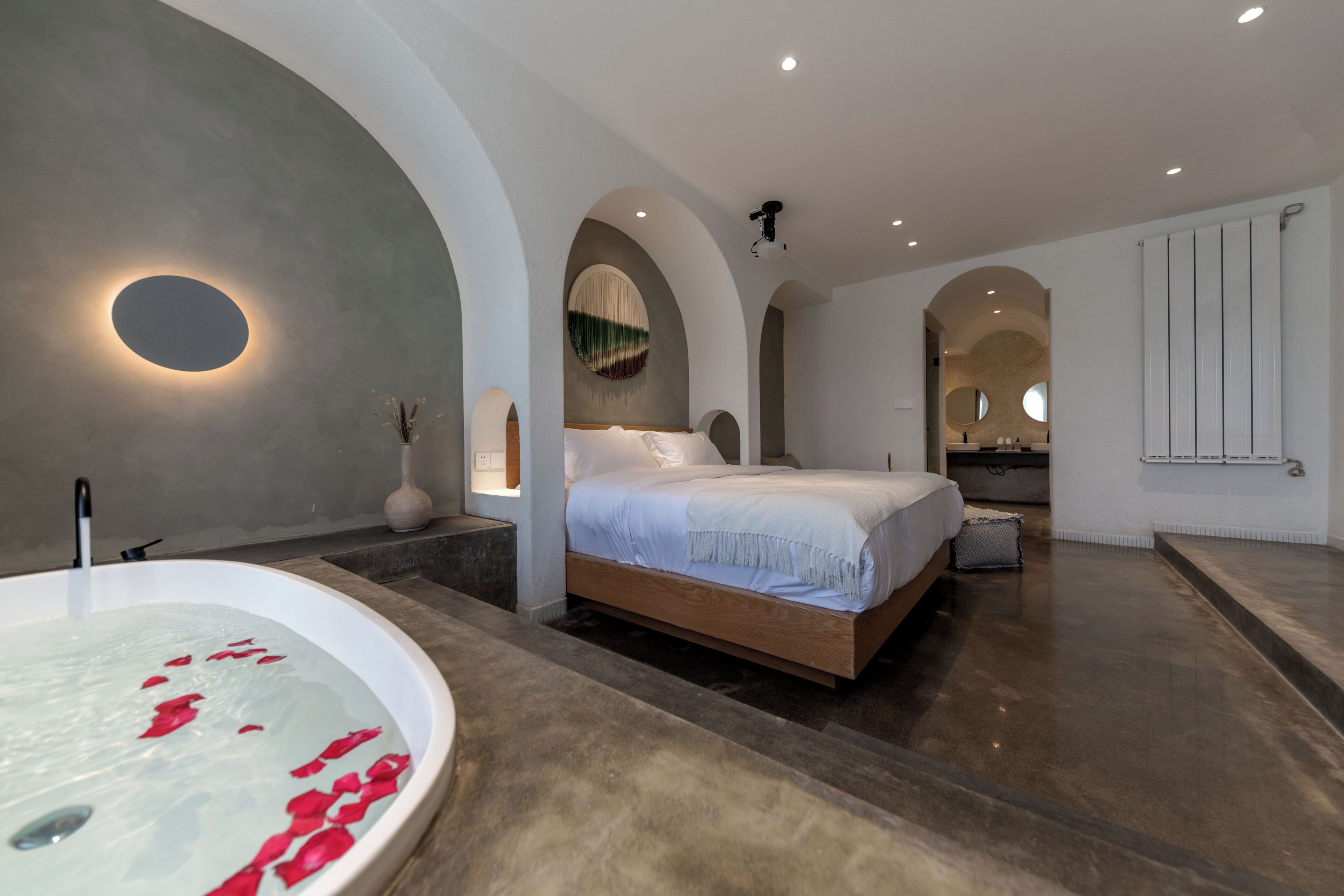 Une chambre moderne avec un grand lit, une baignoire remplie de pétales de roses et une salle de bain dotée d’un miroir et d’un radiateur.