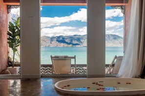 A room with a view of the sea, a bathtub with rose petals, and a chair.