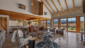 A spacious living area with a high ceiling, wooden beams, a large window offering a mountain view, and a cozy seating arrangement. - Toi Point Lodge - A rural luxury villa inspired by the charm of a historic stone farm building. (Queenstown)