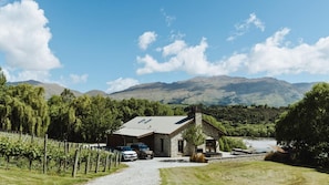 A stone building with a sloped roof, a gravel driveway, parked cars, and a vineyard. - Toi Point Lodge - A rural luxury villa inspired by the charm of a historic stone farm building. (Queenstown)