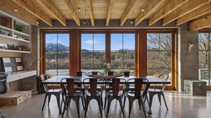 A dining area with a large table and chairs, large windows with a mountain view, wooden ceiling beams, and a stone wall. - Toi Point Lodge - A rural luxury villa inspired by the charm of a historic stone farm building. (Queenstown)