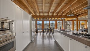 A modern kitchen with a central island, stainless steel appliances, and large windows offering a view of the outdoors. - Toi Point Lodge - A rural luxury villa inspired by the charm of a historic stone farm building. (Queenstown)