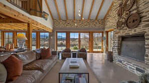 A spacious living room with a stone fireplace, leather sofa, and large windows offering a mountain view. - Toi Point Lodge - A rural luxury villa inspired by the charm of a historic stone farm building. (Queenstown)