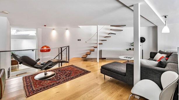 Een moderne woonkamer met een zwarte leren fauteuil, een grijze bank, een rood-zwart tapijt, een trap en een wit plafond.
