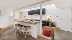 Een moderne keuken met een centraal eiland, barkrukken en een rieten stoel met een rode kussenzitting.