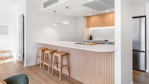 A modern kitchen with a white countertop, wooden stools, stainless steel appliances, and a wooden ceiling vent. - Modern Coastal Escape at Swell 2028 (Gold Coast)