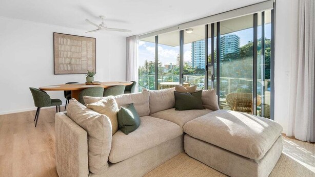 A modern living room with a sectional sofa, a dining table with chairs, and a large window with a view of buildings. - Modern Coastal Escape at Swell 2028 (Gold Coast)