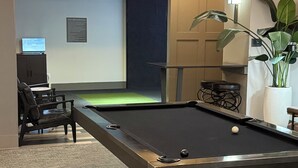 Uma mesa de bilhar com uma bola de sinuca, uma televisão sobre um móvel, uma planta e uma porta que dá para outro quarto.