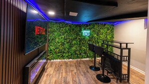 Une pièce avec un plancher en bois, un écran de télévision affichant le logo « NETFLIX », un mur vert orné de l’inscription « RÊVEZ GRAND », et un coin bar avec des chaises.