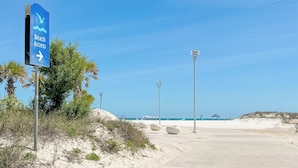 Ein Hinweisschild für den Strandzugang, ein Betonweg und ein Strandabschnitt mit Palmen.
