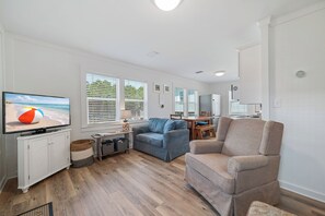 Uma sala de estar com televisão, sofá, poltrona, mesa de jantar e área de cozinha.