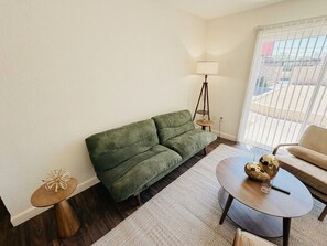 Een woonkamer met een groene bank, een ronde salontafel met decoratieve items, een vloerlamp en uitzicht door een raam met jaloezieën.