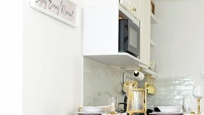 Een keuken met witte kastjes, een magnetron, een wit aanrecht en twee zwarte stoelen met gouden poten.