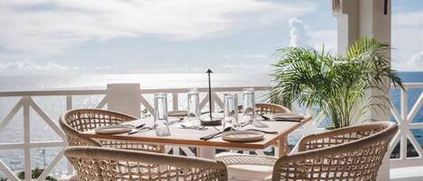 A dining area with wicker chairs and a table set for two, overlooking a scenic ocean view.