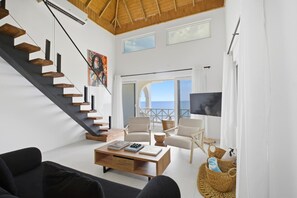 A modern living room with a staircase, a flat-screen TV, and a view of the sea. - 1BR Modern Oceanfront Loft w/ Kitchenette - Syd’s Too (Negril)