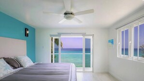 A bedroom with a bed, a ceiling fan, a view of the ocean through a sliding door, and a window with a view of the ocean. - Tideline- 4BR Beachfront Modern Villa (St. James)