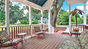 A covered outdoor area with wooden flooring, a seating area with chairs and a sofa, and a view of a house and trees.