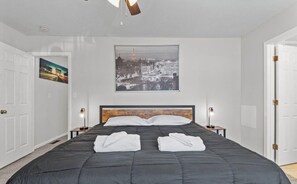 Ein Hotelzimmer mit einem großen Bett, zwei Nachttischen mit Lampen, einem Stadtbild an der Wand und einer Tür links.